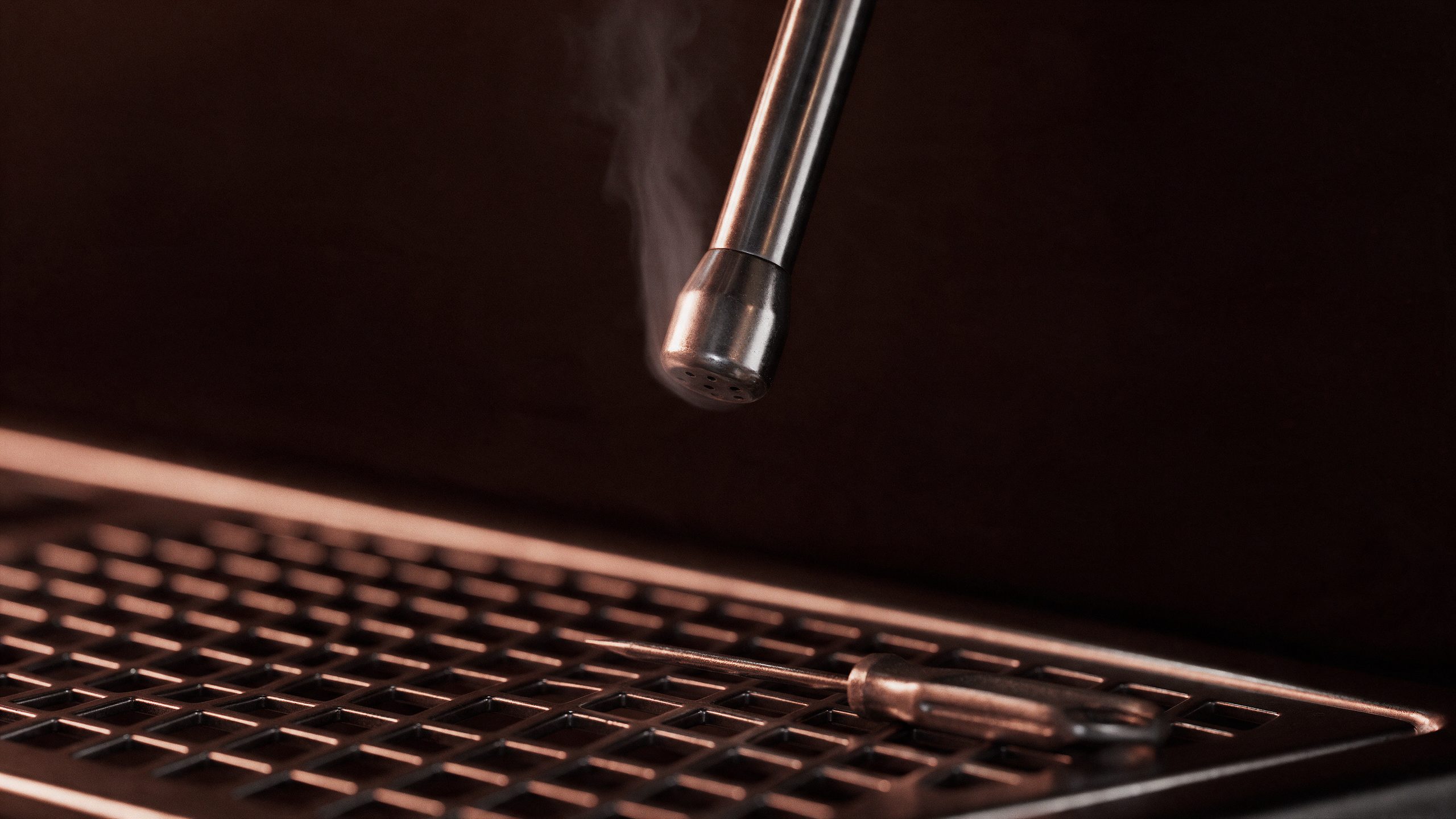 Espresso machine steam wand tip releasing a wisp of steam above a drip tray with a cleaning pin tool